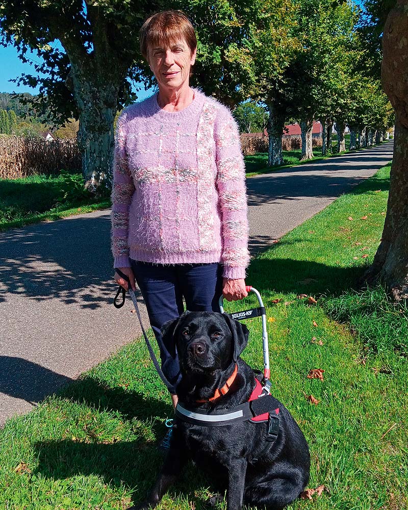 Josiane Chaloyard et Odéon, labrador noir, lors de la remise. La photo est prise sur le site de l'école à Misérieux, Odéon est assis au côté de sa nouvelle maîtresse qui tient son harnais.