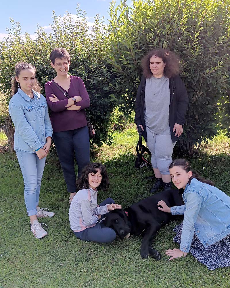 Roseline Souton lors de la remise de Skittle, labrador noir allongé dans l'herbe. 3 adolescentes et une adulte l'entourent, deux des jeunes filles sont assises à côté du chien et lui prodiguent quelques caresses.