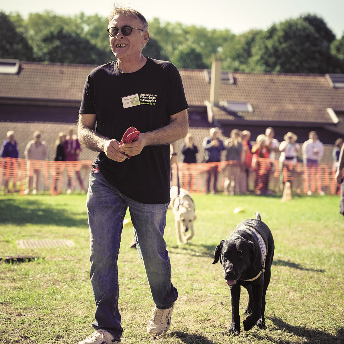 Martial Herrscher en pleine action, en démonstration à l'occasion d'une journée portes ouvertes de l'association. Il évolue dans l'herbe, accompagné d'un élève chien guide noir. A l'arrière-plan, les spectateurs massés derrière les barrières de sécurité.
