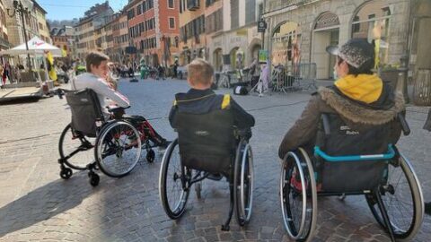 Trois personnes en fauteuil roulant discutent dans le centre piéton de Chambéry. Il fait un soleil d'hiver.