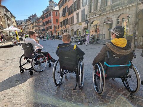 Trois personnes en fauteuil roulant discutent dans le centre piéton de Chambéry. Il fait un soleil d'hiver.