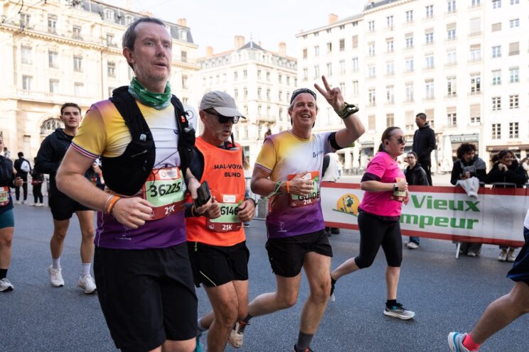 Un coureur malvoyant portant un dossard orange court entouré de ses deux guides vêtus de tee-shirts violet et jaune. L’un des guides sourit et fait un signe de la paix avec la main. Ils avancent dans une rue du centre-ville, au milieu d’autres participants et sous le regard de spectateurs derrière des barrières.