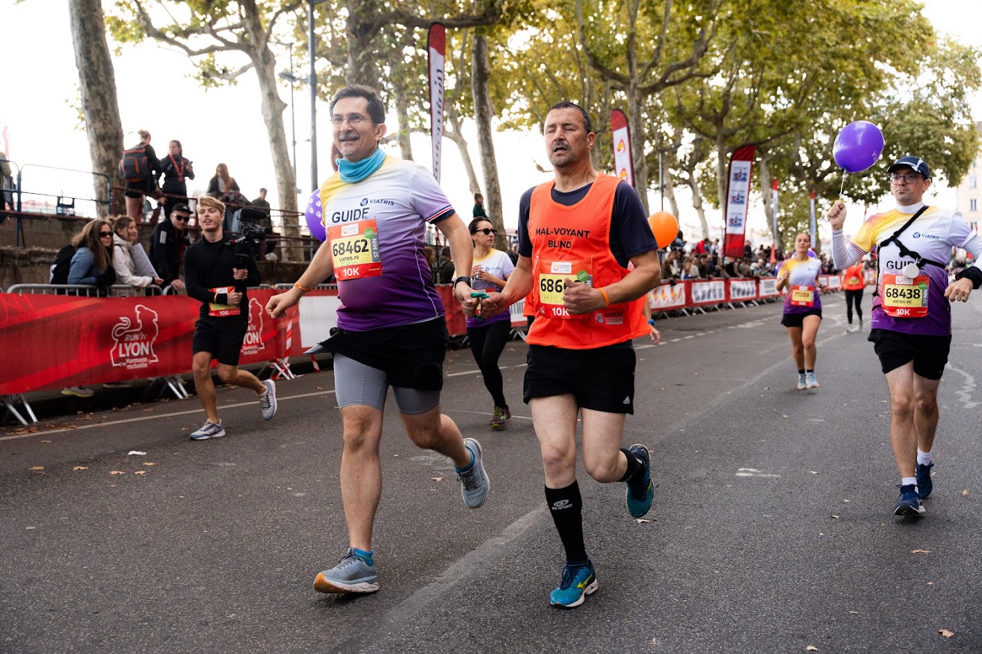 Un guide court aux côtés d’un participant malvoyant portant un dossard orange. Ils avancent en se tenant par une corde de guidage, entourés d’autres coureurs sur une avenue bordée d’arbres. Des spectateurs encouragent derrière les barrières rouges de l’événement “Run In Lyon”.