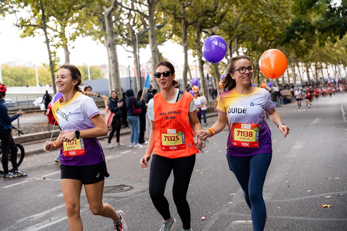 Deux guides courent aux côtés d’une participante malvoyante portant un dossard orange. Elles avancent en se tenant par le poignet, souriantes, sur une avenue bordée d’arbres. Les guides portent des tee-shirts colorés et tiennent des ballons violets et orange. Des spectateurs applaudissent en arrière-plan.