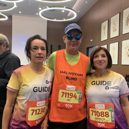Deux guides et une coureuse malvoyante posent côte à côte dans une salle. La coureuse porte un dossard orange avec l’inscription « Mal-voyant / Blind » et des lunettes teintées. Les deux guides, une femme à gauche et une femme à droite, portent des tee-shirts de course aux couleurs violette et jaune avec le mot « Guide ». Tous trois sourient légèrement en direction de l’appareil photo.