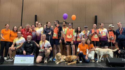 Un groupe de coureurs et de bénévoles pose sur une scène avec plusieurs chiens guides. Certains portent des médailles et des dossards colorés. L’ambiance est joyeuse et un grand chèque au nom de l’association Chiens Guides est présenté au premier plan.