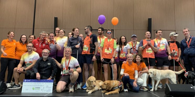 Un groupe de coureurs et de bénévoles pose sur une scène avec plusieurs chiens guides. Certains portent des médailles et des dossards colorés. L’ambiance est joyeuse et un grand chèque au nom de l’association Chiens Guides est présenté au premier plan.