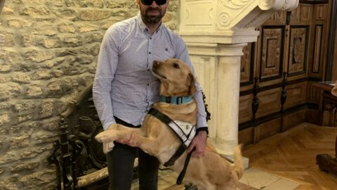 Un homme portant des lunettes de soleil se tient debout dans une pièce aux murs en pierre et au décor ancien. À ses côtés, un chien guide labrador sable, debout sur ses pattes arrière, pose ses pattes avant sur lui comme pour un câlin. L’homme sourit légèrement en tenant le chien par le harnais.