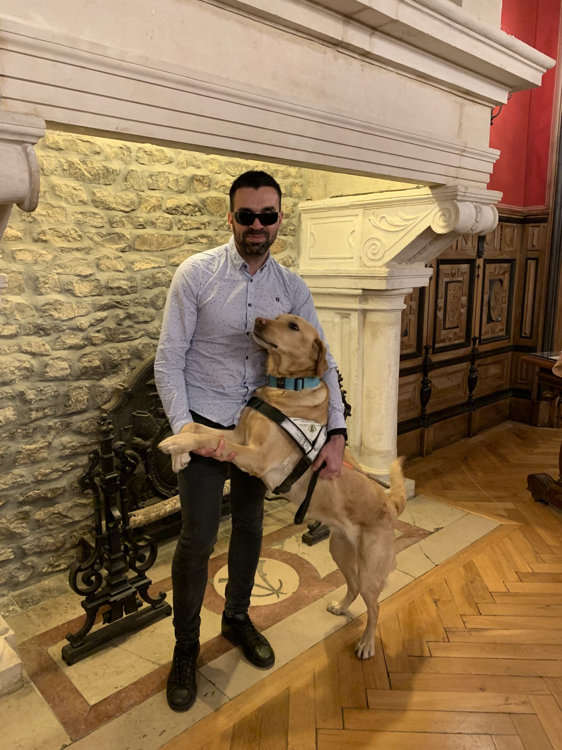 Un homme portant des lunettes de soleil se tient debout dans une pièce aux murs en pierre et au décor ancien. À ses côtés, un chien guide labrador sable, debout sur ses pattes arrière, pose ses pattes avant sur lui comme pour un câlin. L’homme sourit légèrement en tenant le chien par le harnais.