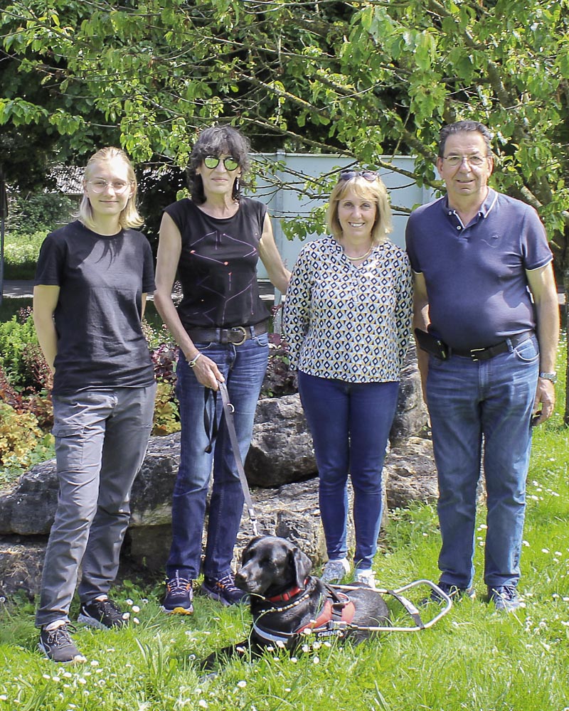 Isabelle et Upsy lors de la remise à Misérieux, devant un massif de rocailles, entourés de végétation. Upsy est couché aux pieds d'Isabelle, équipé de son harnais, ils sont tous deux entourés d'Éloïse Guignard et Françoise et Bertrand Charles.