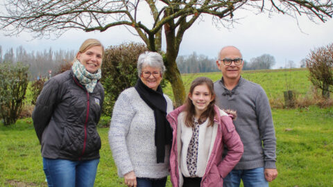 Deux femmes, dont l'une porte une doudoune noire avec le logo de l'association de chiens guides de lyon, un homme et une jeune fille sont tous les 4 debout sur un trottoir. Assis devant eux et regardant la jeune fille, se tient un labrador noir avec un collier bleu. En arrière plan un arbre sans feuilles et un pré vert