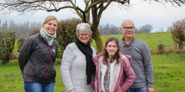 Deux femmes, dont l'une porte une doudoune noire avec le logo de l'association de chiens guides de lyon, un homme et une jeune fille sont tous les 4 debout sur un trottoir. Assis devant eux et regardant la jeune fille, se tient un labrador noir avec un collier bleu. En arrière plan un arbre sans feuilles et un pré vert