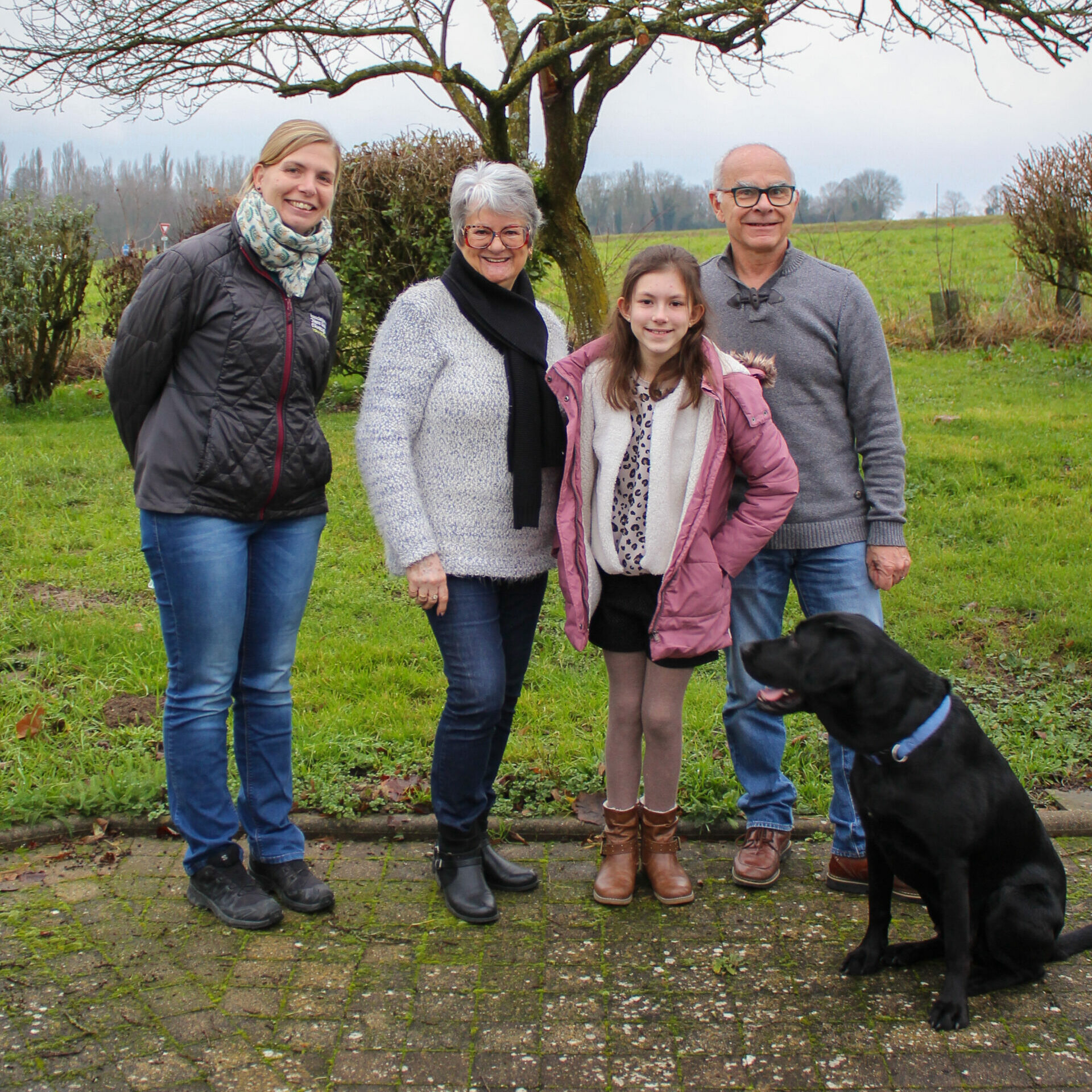 Deux femmes, dont l'une porte une doudoune noire avec le logo de l'association de chiens guides de lyon, un homme et une jeune fille sont tous les 4 debout sur un trottoir. Assis devant eux et regardant la jeune fille, se tient un labrador noir avec un collier bleu. En arrière plan un arbre sans feuilles et un pré vert