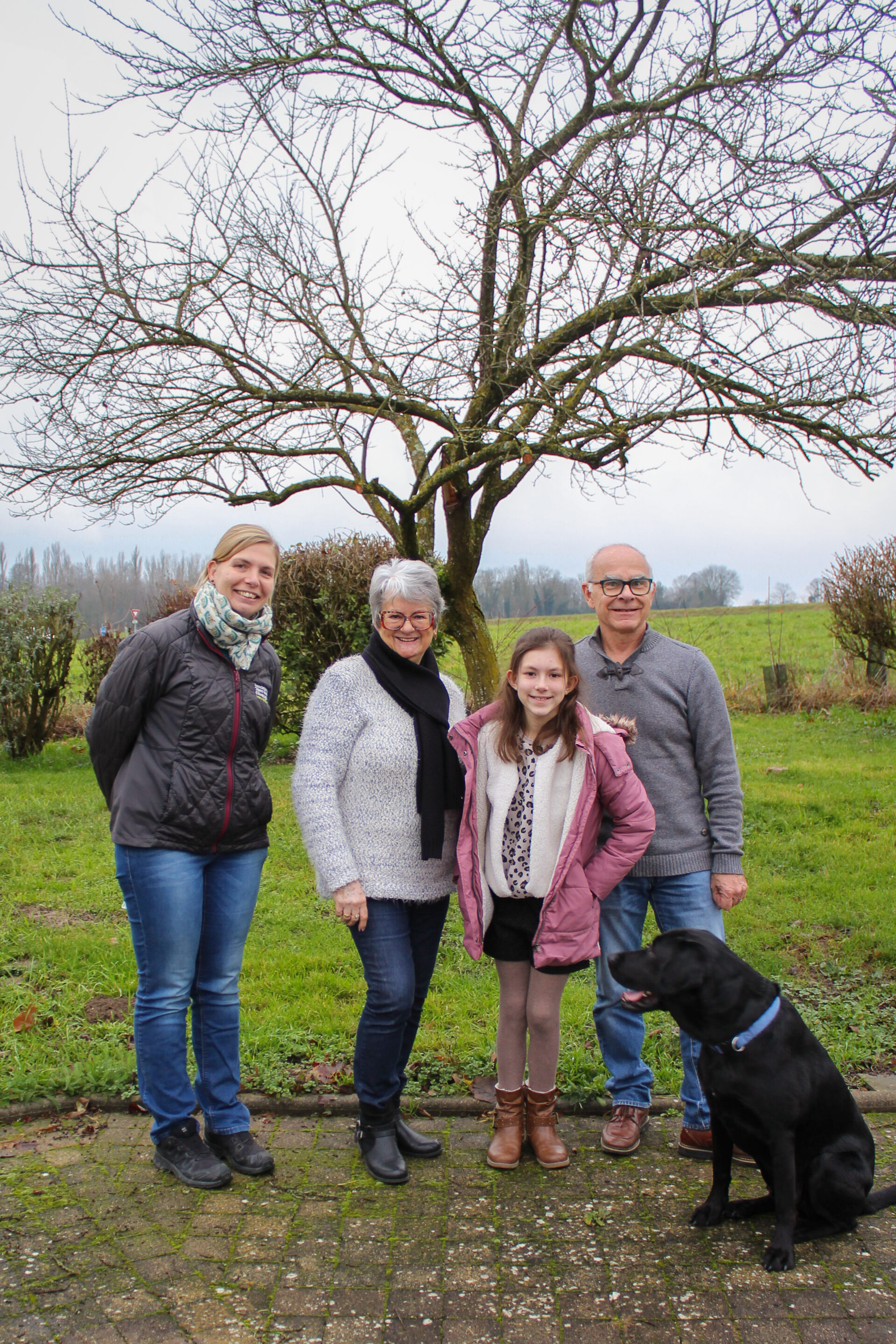 Deux femmes, dont l'une porte une doudoune noire avec le logo de l'association de chiens guides de lyon, un homme et une jeune fille sont tous les 4 debout sur un trottoir. Assis devant eux et regardant la jeune fille, se tient un labrador noir avec un collier bleu. En arrière plan un arbre sans feuilles et un pré vert