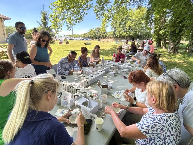 Les familles d'accueil, maîtres de chiens guides, salariés et familles d'accueil peignent leur tasse en céramique sur une grande table installée dans l'herbe, à l'ombre des arbres, avec un beau ciel bleu en arrière-plan.