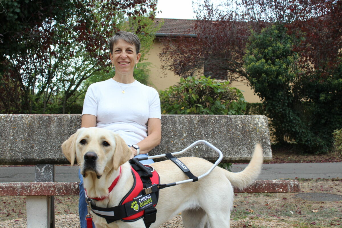 Annie, souriante, assise sur un banc en pierre dans un parc, pose la main sur Venga, jeune labrador sable équipée d'un harnais de chien guide lors de sa remise.