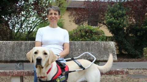 Annie, souriante, assise sur un banc en pierre dans un parc, pose la main sur Venga, jeune labrador sable équipée d'un harnais de chien guide lors de sa remise.