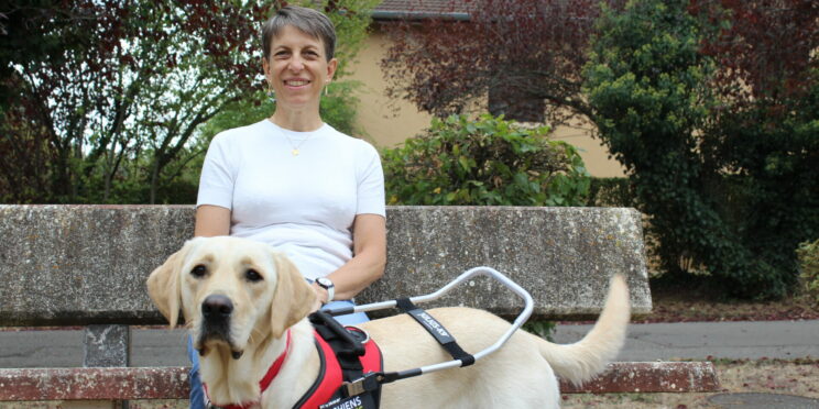 Annie, souriante, assise sur un banc en pierre dans un parc, pose la main sur Venga, jeune labrador sable équipée d'un harnais de chien guide lors de sa remise.