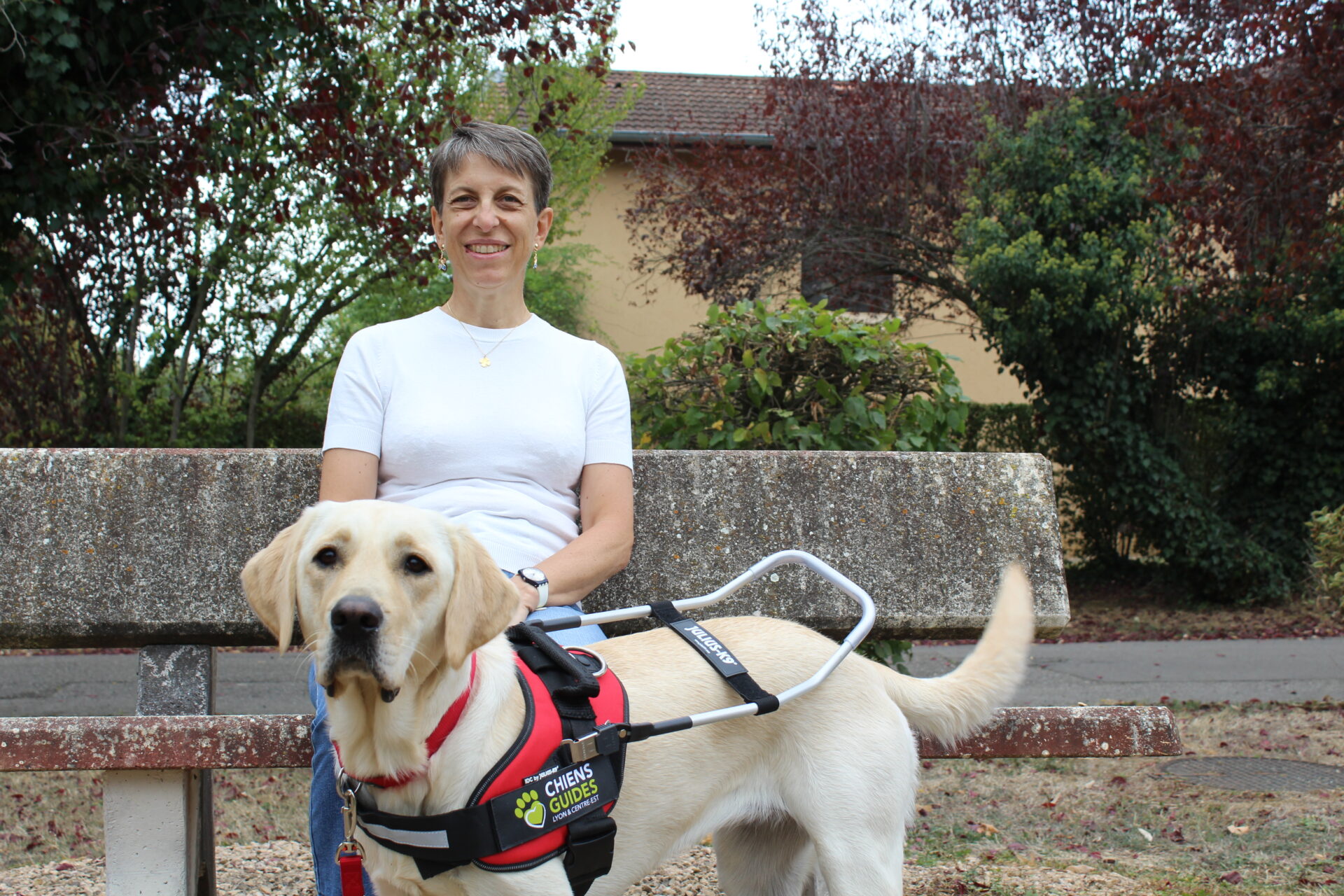 Annie, souriante, assise sur un banc en pierre dans un parc, pose la main sur Venga, jeune labrador sable équipée d'un harnais de chien guide lors de sa remise.