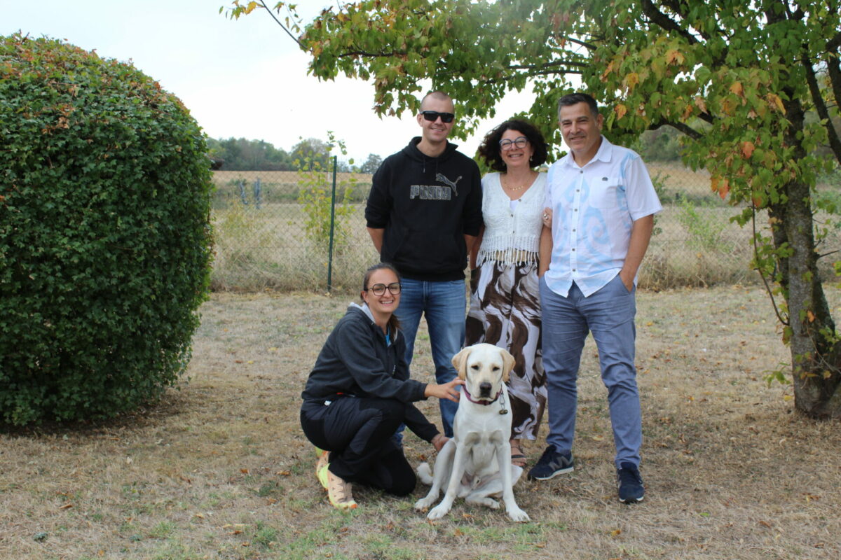Virgin, labrador sable, assis dans un jardin, entouré de quatre personnes souriantes pour sa remise. Accroupie à sa gauche, Laura, notre éducatrice, le caresse. Derrière, debout, Christopher, son nouveau maître aux côtés de la famille Thobois, sous un arbre.