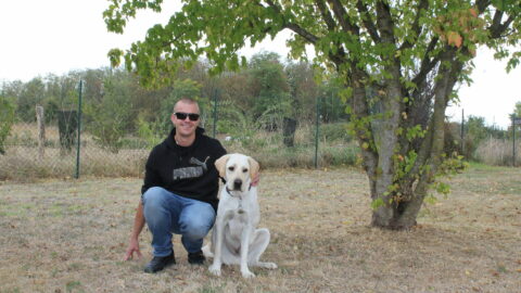 Christopher, souriant en sweat noir et lunettes de soleil, accroupi dans l'herbe, un bras autour de Virgin, son labrador sable assis à ses côtés. Ils posent sous un arbre, devant une clôture grillagée.