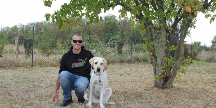 Christopher, souriant en sweat noir et lunettes de soleil, accroupi dans l'herbe, un bras autour de Virgin, son labrador sable assis à ses côtés. Ils posent sous un arbre, devant une clôture grillagée.